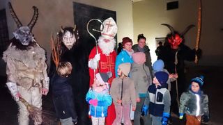 A group of children and adults are standing in front of a person dressed as Santa Claus, a devil, and a bishop. The children are wearing winter clothes and some are wearing hats. The person dressed as the devil is holding a stick with a cross on top.