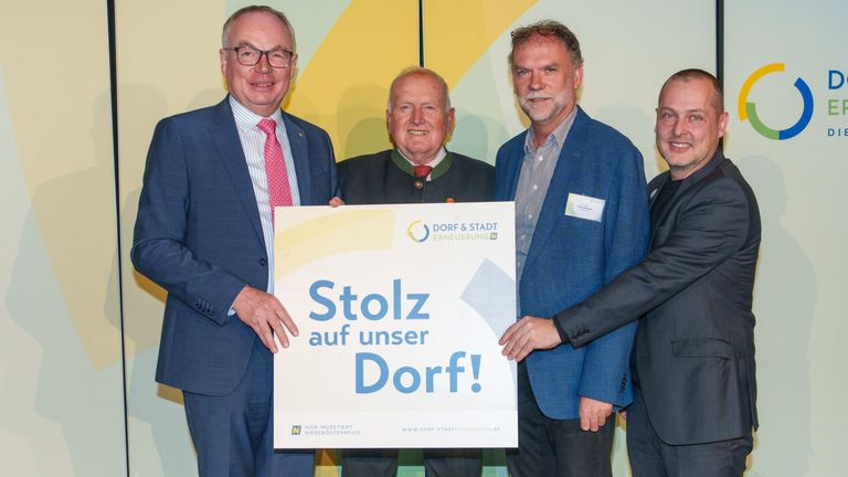 Vier Männer stehen neben einem großen Schild und lächeln. Das Schild zeigt Stolz auf unser Dorf! in fetten blauen Buchstaben. Die Männer sind in Anzügen gekleidet.
