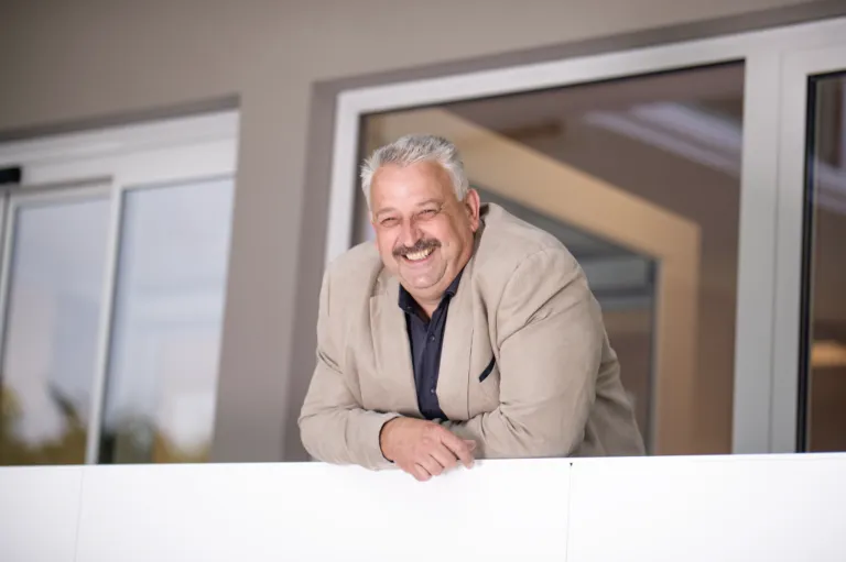 Ein älterer Mann mit einem Lächeln im Gesicht lehnt sich an eine weiße Wand, möglicherweise für ein Foto posierend.