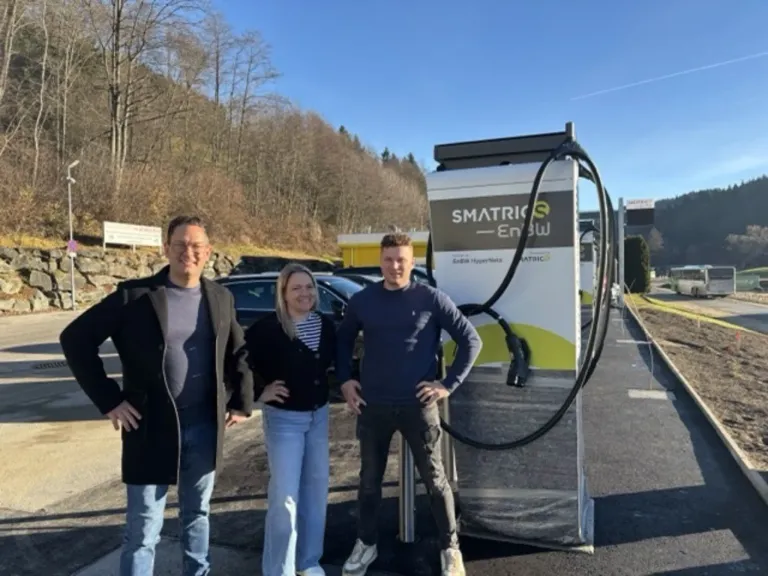 Drei Personen posieren vor einer Ladestation für Elektrofahrzeuge. Dahinter sind Autos in einem Parkplatz abgestellt. Im Hintergrund sind Bäume und ein entferntes Gebäude zu sehen.