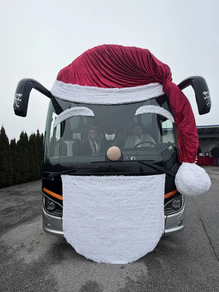 Ein dekorierter Bus mit einem Weihnachtsmannhut, einer Nase und einem Bart ist draußen geparkt. Zwei Personen sitzen im Inneren, einer fährt und der andere lächelt. Bäume und ein Gebäude sind im Hintergrund.