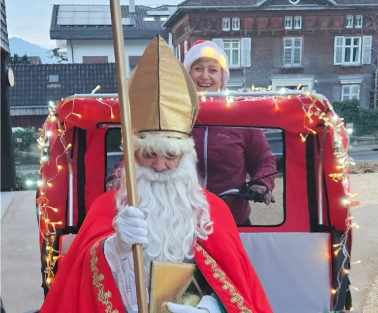 Eine Person, die als Weihnachtsmann gekleidet ist, sitzt in einem dekorierten roten Fahrzeug und lächelt, neben ihr eine Frau in einer Weihnachtsmütze.