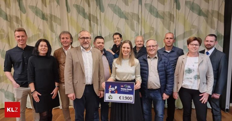 Eine Gruppe von Menschen steht lächelnd für ein Foto. Eine Frau hält ein Schild mit 1.900 €. Hinter ihnen ist ein Vorhang mit grünen Blättern zu sehen.