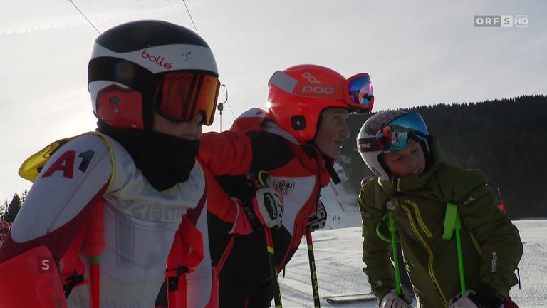 Drei Skifahrer in Helmen stehen auf einem schneebedeckten Feld. Sie tragen Skiausrüstung und halten Skistöcke. Der Himmel ist klar.