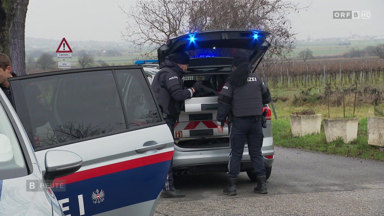 Two police officers are standing next to a police car with its trunk open on a gravel road. One officer is opening the trunk while the other is standing by.