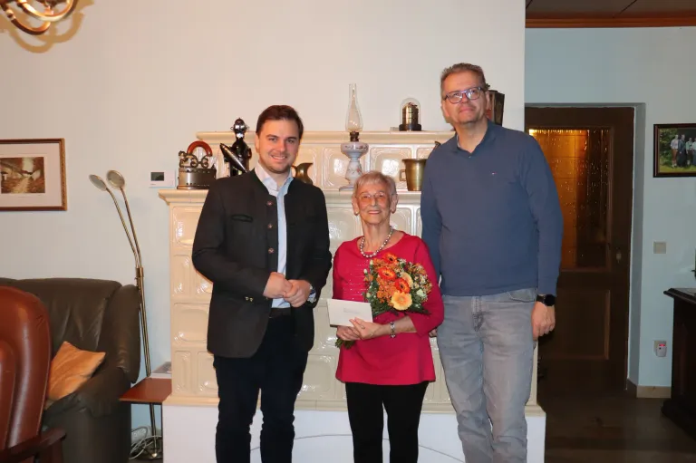 Drei Menschen stehen lächelnd vor einem Kamin. Eine Frau hält einen Blumenstrauß und ein Papier, während der Mann auf der linken Seite ein Papier hält. Dahinter steht ein weißer Kamin mit dekorativen Gegenständen.