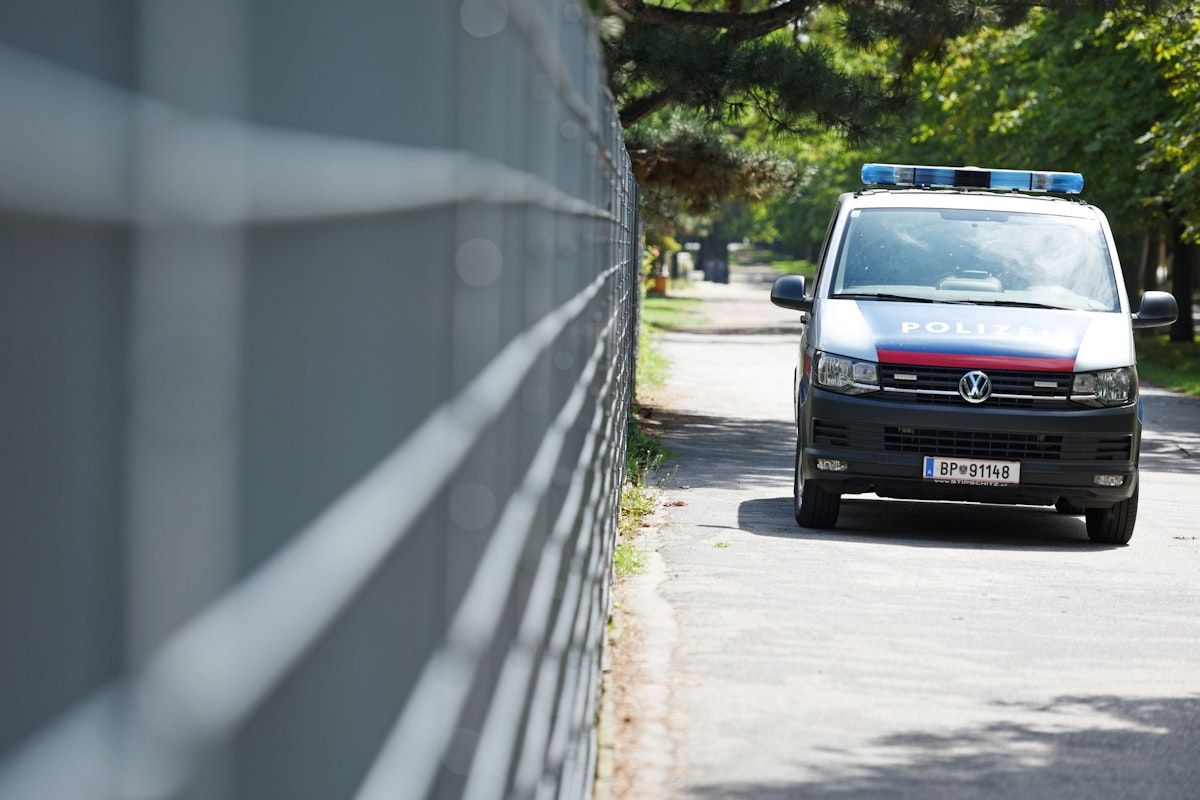 Ein Polizeifahrzeug ist am Straßenrand in der Nähe eines Zauns geparkt. Das Fahrzeug hat ein Nummernschild mit der Aufschrift BP 9114. Die Szene wird von Bäumen beschattet.