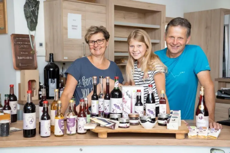 Eine Familie steht lächelnd in einer Küche. Sie stehen vor einem Tisch mit Flaschen und Gläsern. Die Frau trägt eine Brille und ein blaues T-Shirt. Der Mann trägt ein blaues T-Shirt und das Mädchen ein gestreiftes T-Shirt. Hinter ihnen befinden sich Holzschränke.