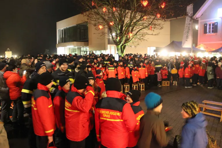 Eine große Menschenmenge, die rote Jacken mit gelben Streifen und schwarze Mützen trägt, hat sich um einen mit roten Lichtern geschmückten Baum versammelt. Sie stehen vor einem Gebäude mit großen Fenstern.