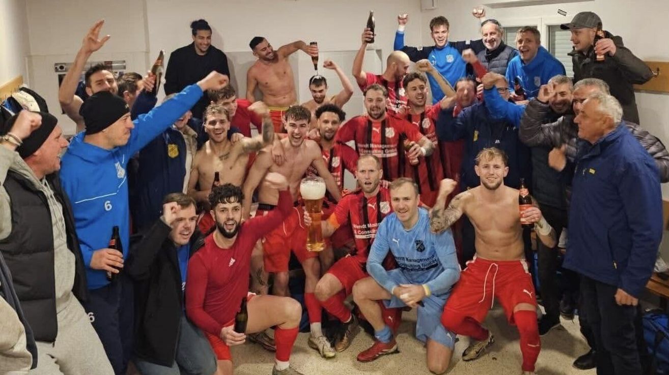 A team of soccer players in red and blue uniforms celebrate a victory. They hold bottles and cups of beer.