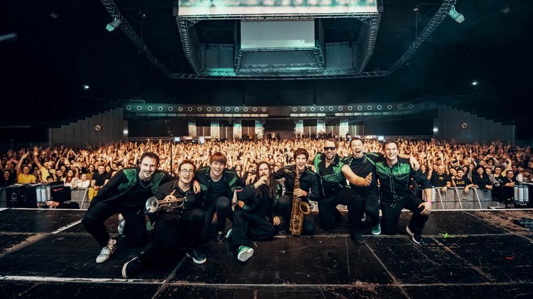 Eine Band posiert für ein Foto auf der Bühne vor einem jubelnden Publikum. Sie tragen alle passende grüne und schwarze Kleidung.