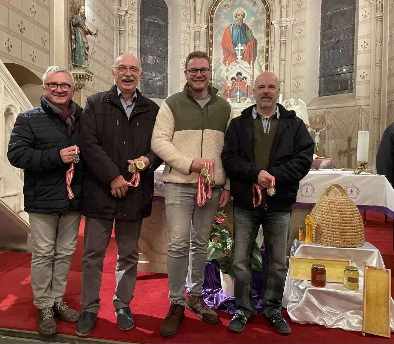 Vier Männer in Jacken stehen in einer Kirche. Sie halten goldene Medaillen. Hinter ihnen steht ein Altar mit einem Korb, Kerzen und Blumen.