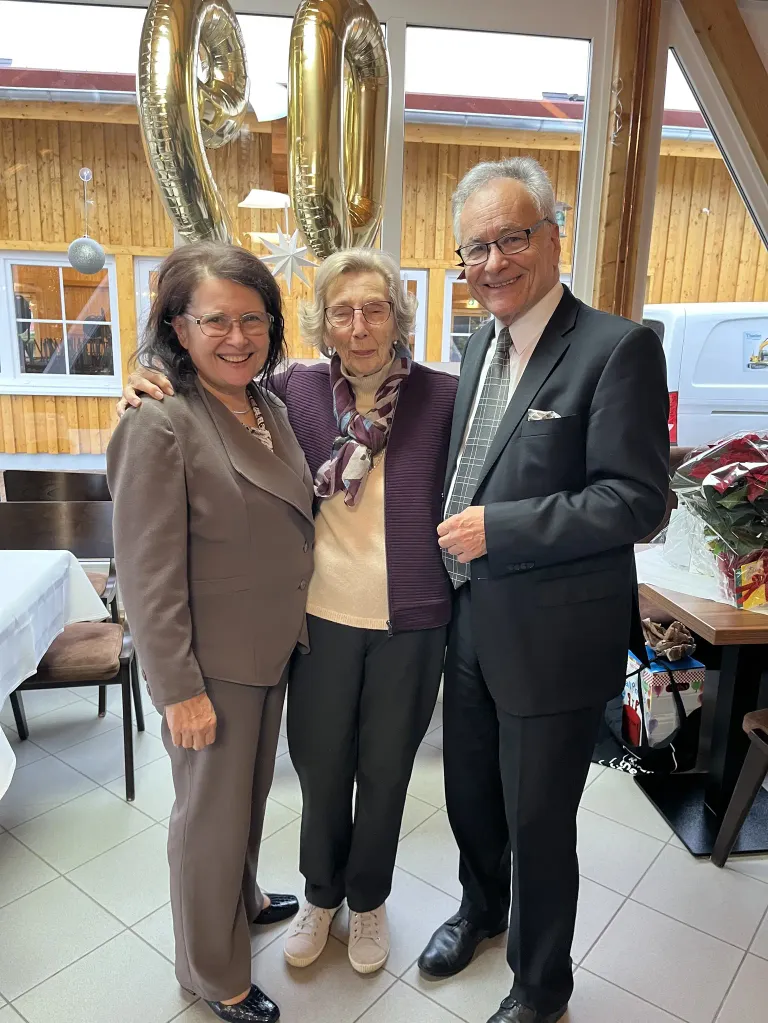 Drei Menschen in formeller Kleidung stehen in einem Raum mit Holzwänden. Ein Mann auf der rechten Seite trägt eine Brille und eine Krawatte. Die Frau in der Mitte trägt einen Schal. Hinter ihnen steht ein Tisch mit einer Blumendekoration.