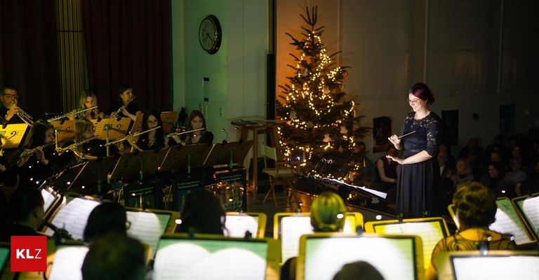 Eine Frau dirigiert eine Band in einem Raum mit Musikinstrumenten und einem Weihnachtsbaum. Menschen sitzen auf Stühlen und spielen Instrumente. An der Wand hängen eine Uhr und ein Bilderrahmen.