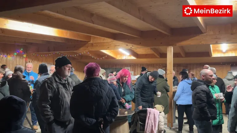 Eine Gruppe von Menschen ist drinnen versammelt, wahrscheinlich bei einer Gemeinschaftsveranstaltung. Einige tragen Winterkleidung, und eine Person mit rosa Haaren sticht hervor. Eine Holzdecke und warmes Licht schaffen eine gemütliche Atmosphäre.