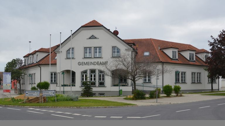 Ein Gemeindeamt mit rotem Dach, weißen Wänden und großen Fenstern ist zu sehen. Das Gebäude heißt 'GEMEINDEAM'. Es hat einen kleinen Garten vor dem Gebäude mit einem Schild und Straßenlaternen.