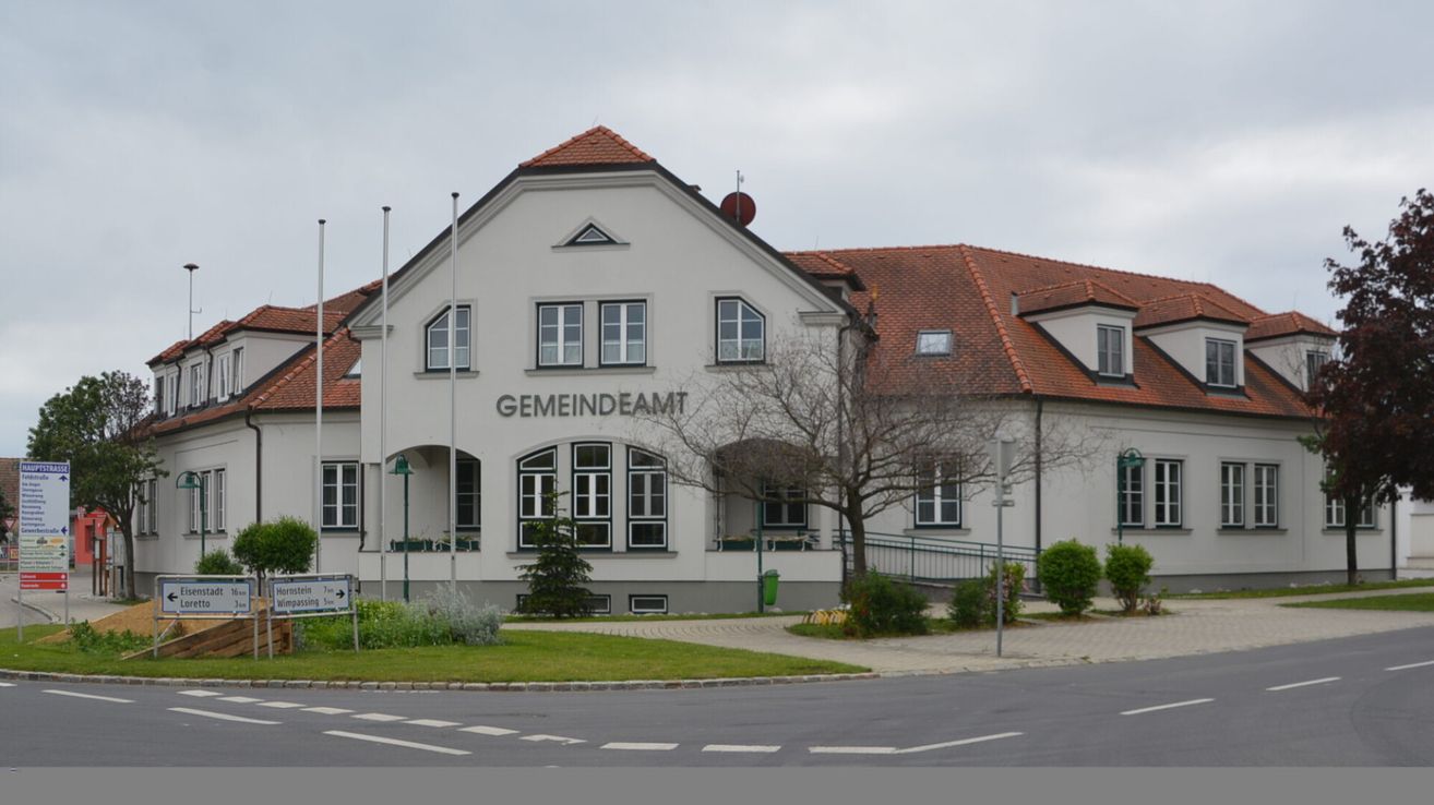 Ein Gemeindeamt mit rotem Dach, weißen Wänden und großen Fenstern ist zu sehen. Das Gebäude heißt 'GEMEINDEAM'. Es hat einen kleinen Garten vor dem Gebäude mit einem Schild und Straßenlaternen.