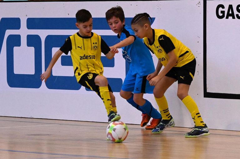 Drei junge Jungen spielen Hallenfußball. Zwei sind in Gelb und einer in Blau. Sie jagen einem Fußball hinterher.