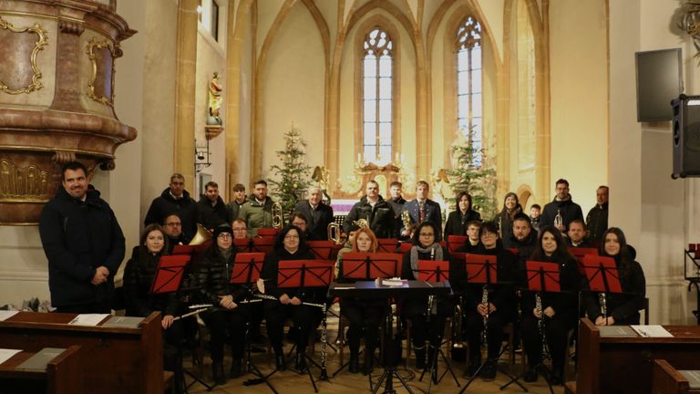 Eine Gruppe von Musikern posiert für ein Foto in einer Kirche, trägt Winterkleidung und hält Musikinstrumente. Dahinter stehen zwei Weihnachtsbäume, und die Kirche hat Bogenfenster und eine Statue.