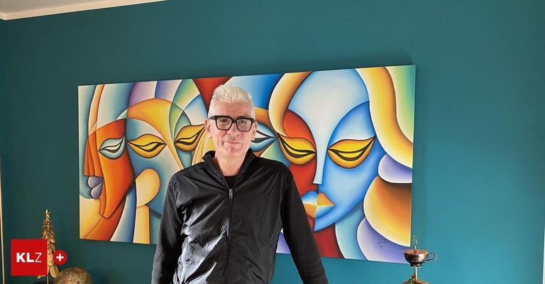 Ein Mann mit weißen Haaren steht vor einem großen abstrakten Gemälde an einer türkisen Wand, trägt eine schwarze Jacke und eine Brille.