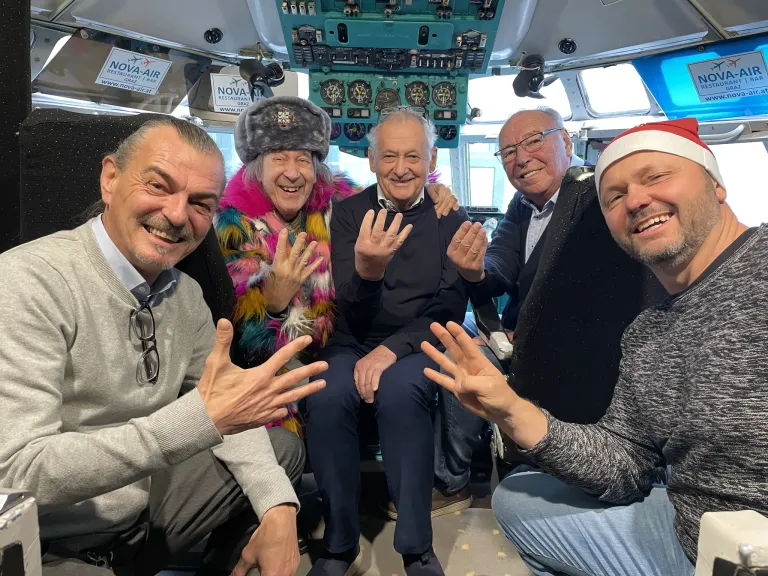 Fünf Menschen sitzen in einer Flugzeugkabine, lächeln und posieren für ein Foto. Eine Frau in bunter Kleidung steht zwischen zwei Männern. Dahinter befindet sich ein Kontrollpanel mit verschiedenen Anzeigen und einem Schild.