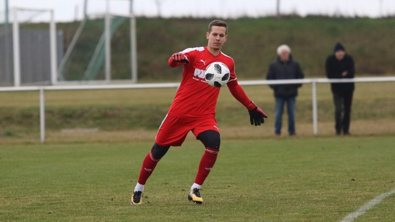 Ein Fußballspieler in roter Uniform kickt einen Fußball auf einem Feld. Ein älterer Mann steht im Hintergrund in der Nähe eines weißen Zauns.