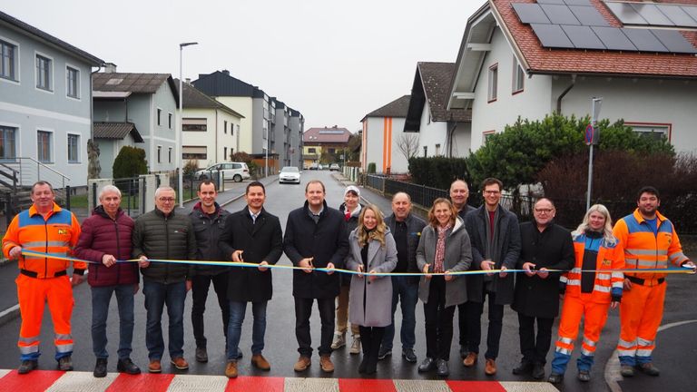 Eine Gruppe von Menschen steht hinter einem Band auf einer Straße und lächelt für ein Foto. Sie sind von Häusern und Autos umgeben.