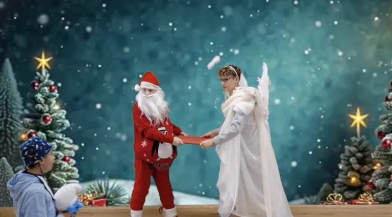 Eine Person als Weihnachtsmann überreicht einem Engel auf einer verschneiten Bühne ein Geschenk.