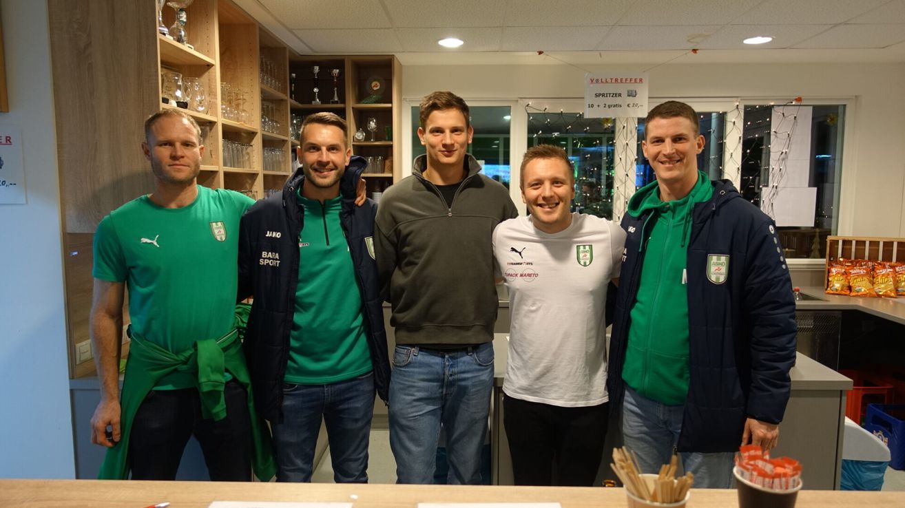 Fünf Männer in Sportkleidung stehen in einem Raum mit einem Holztisch und Regalen gefüllt mit Glaswaren und Trophäen. Der Mann in der Mitte trägt eine graue Kapuzenjacke.