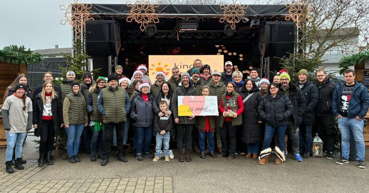 Eine Gruppe von Menschen in Winterkleidung posiert für ein Foto vor einer festlich dekorierten Bühne mit einem Banner. Sie halten einen großen Spendenscheck.