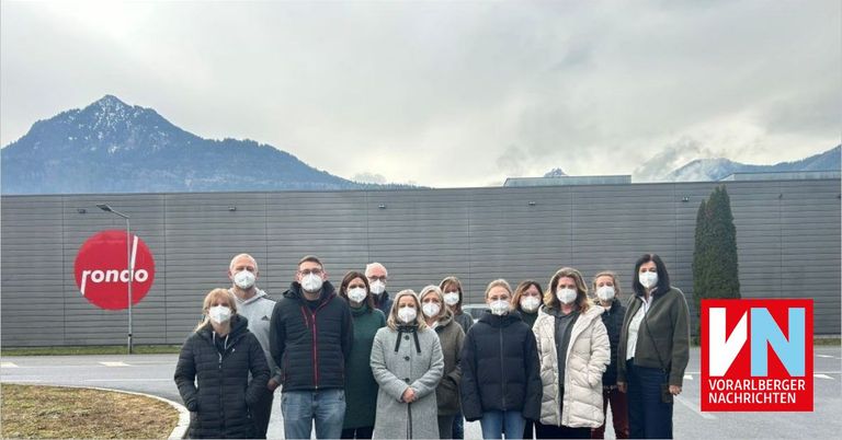 Eine Gruppe von Menschen mit Masken steht vor einem Gebäude mit Bergen im Hintergrund.