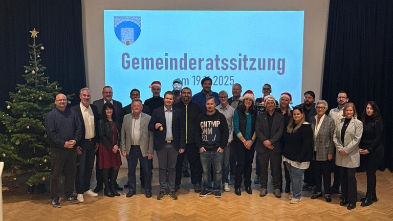 Gruppenfoto bei einer Gemeinderatssitzung am 19. März 2025. Ein großer digitaler Schriftzug im Hintergrund lautet 'Gemeinderatssitzung'. Die Mitglieder tragen festliche Mützen.