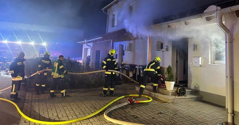 Firefighters are outside a house at night, spraying water and smoke. They are wearing helmets and boots. There is a hose on the ground, and a potted plant on the porch.