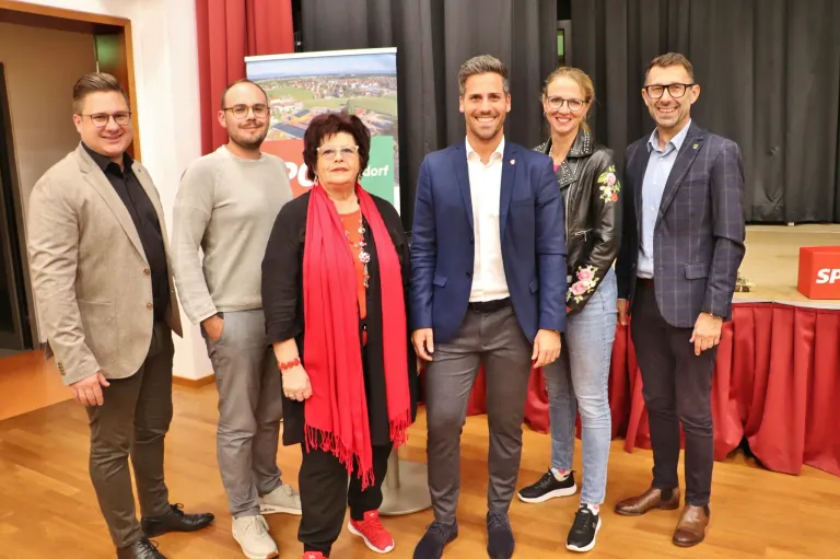 Fünf Personen stehen zusammen, lächeln und posieren für ein Foto. Die Frau in der Mitte trägt einen roten Schal. Dahinter ist ein roter Hintergrund mit einem Schild und einer Stadtlandschaft zu sehen.