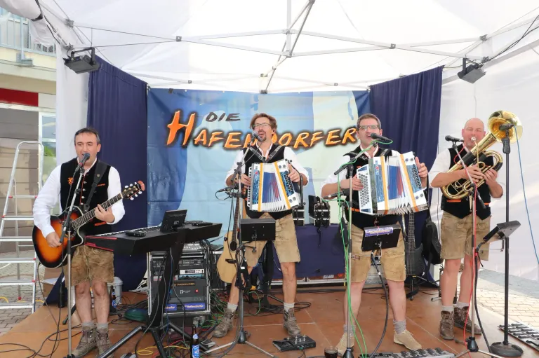 Eine Gruppe von Musikern tritt auf der Bühne auf. Zwei Männer spielen Akkordeons und singen in Mikrofone. Ein weiterer Mann spielt Gitarre. Hinter ihnen steht ein Banner mit der Aufschrift 'DIE HAFENWORFER'. Auf dem Boden befinden sich ein Keyboard und andere Musikinstrumente.