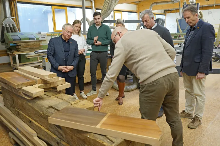 Eine Gruppe von Menschen in einer Werkstatt, die Holzbretter betrachten. Ein älterer Mann zeigt auf ein Stück Holz. Hinter ihm steht ein Mann mit Brille, der spricht.