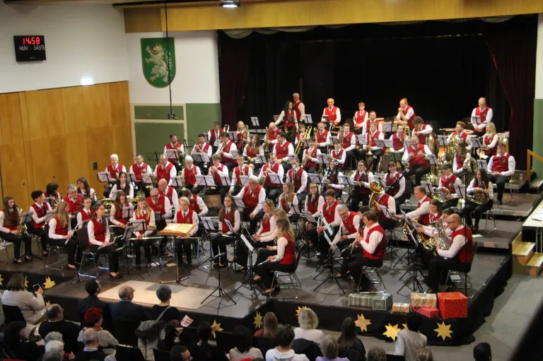 Ein großes Blasorchester spielt auf der Bühne, mit Musikern in roten Westen, die verschiedene Instrumente spielen. Das Publikum sitzt vor der Bühne, und die Bühne ist mit Noten und Geschenkboxen dekoriert.
