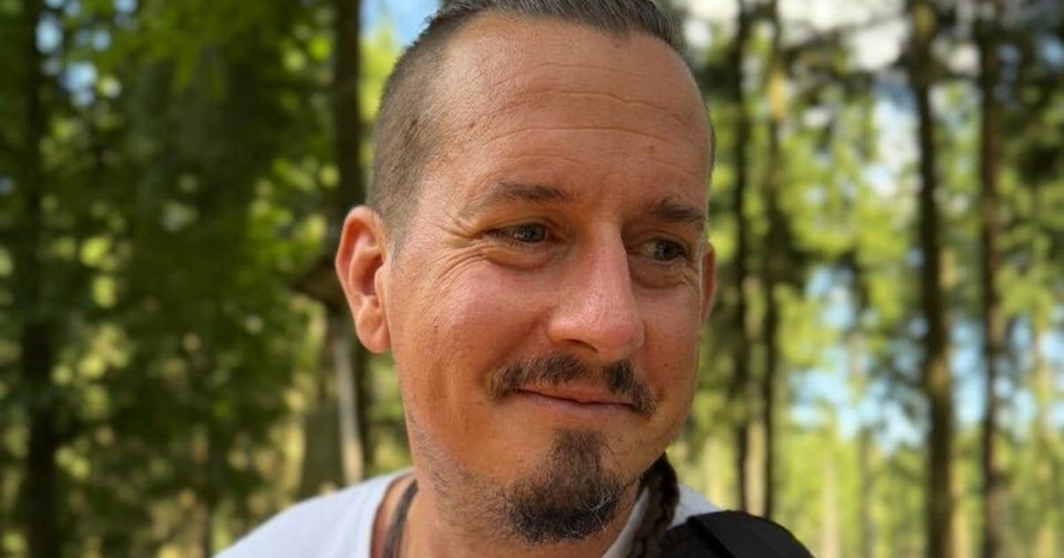 Ein Mann mit Bart und Oberlippenbart steht in einem Wald. Er trägt ein weißes T-Shirt und hat geflochtene Haare. Die Bäume hinter ihm sind verschwommen.