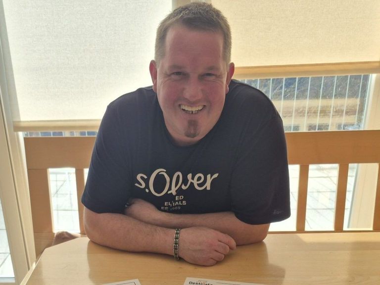 Ein Mann mit einem Schnurrbart lächelt, während er mit verschränkten Armen an einem Tisch sitzt, ein schwarzes T-Shirt mit der Aufschrift 's.Over' trägt.