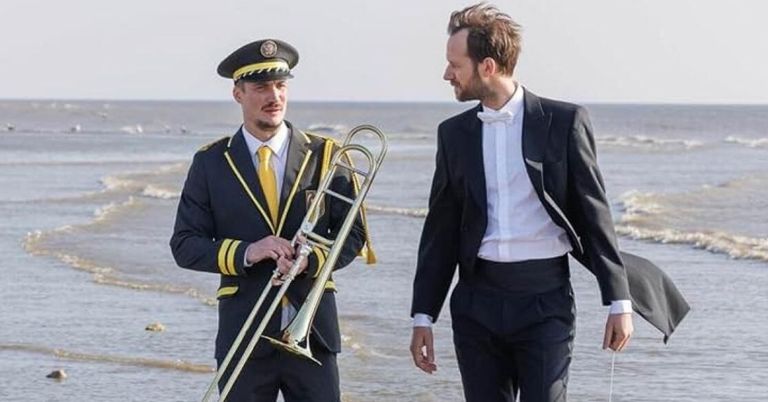 Zwei Männer stehen am Strand, einer hält ein Posaune und beide sind formell gekleidet. Der Mann mit der Posaune trägt eine Uniform mit gelben Streifen.