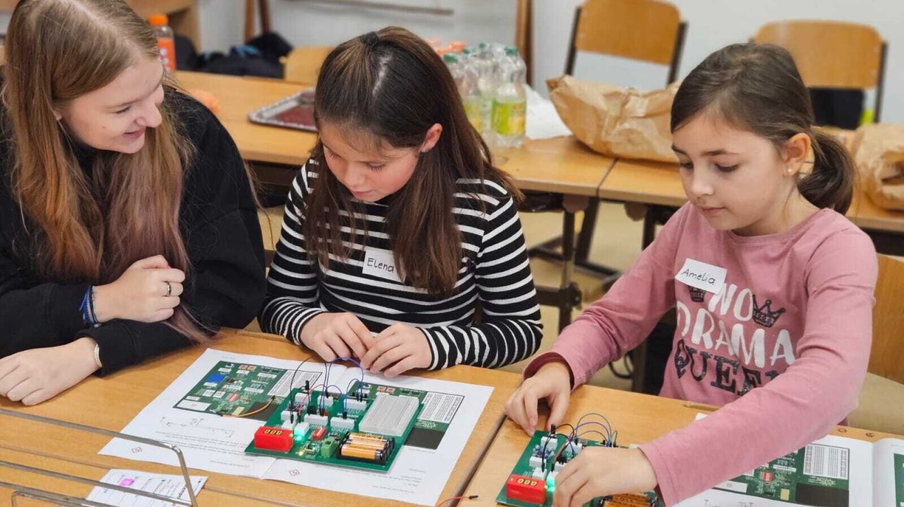 Zwei Mädchen sitzen an einem Schreibtisch und arbeiten an einem Elektronikprojekt. Eines der Mädchen, Elena, konzentriert sich auf eine Leiterplatte. Ein anderes Mädchen in einem rosa Shirt arbeitet neben ihr ebenfalls an einem ähnlichen Projekt.