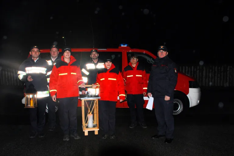Eine Gruppe von Feuerwehrleuten in roten Jacken steht bei Nacht vor einem roten Van und hält eine Kerze. Sie posieren für ein Foto.