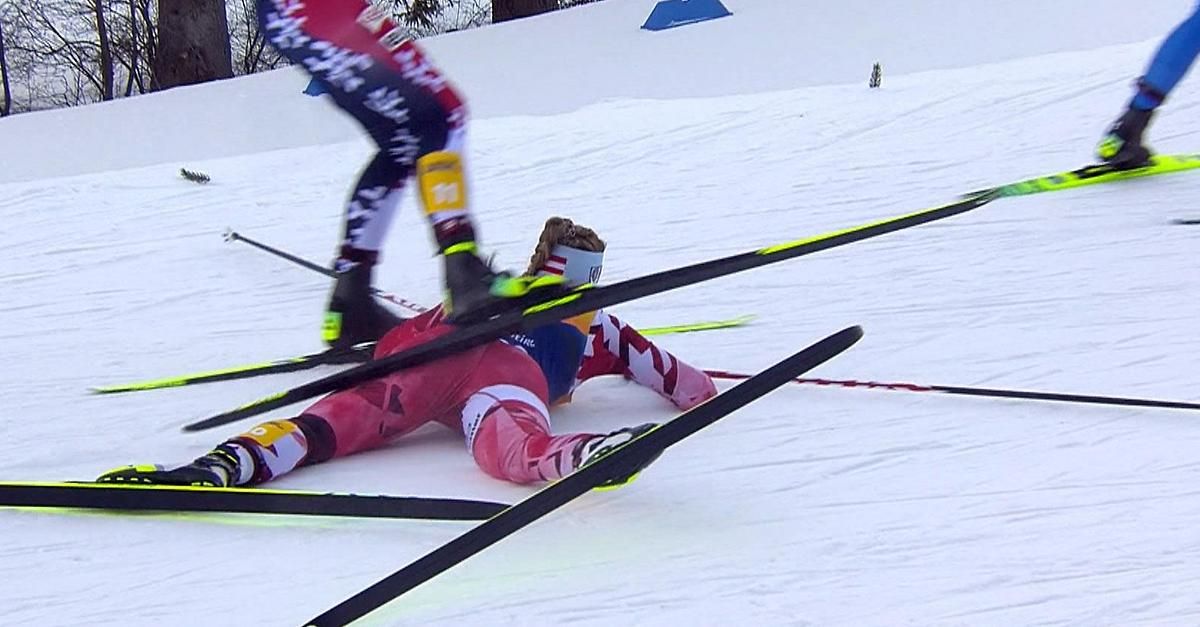 Ein Skifahrer liegt auf dem Schnee mit den Skiern in der Luft, ein anderer Skifahrer steht in der Nähe und trägt ein blau-weißes Trikot.