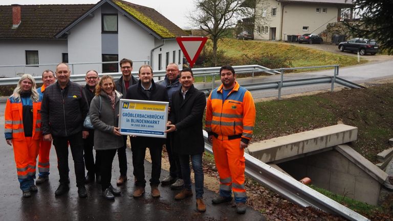 Eine Gruppe von Menschen steht vor einem Gebäude, einer hält ein Schild mit der Aufschrift 'Groblerbachbrucke in Blindenmarkt'. Sie scheinen ein Bauprojekt zu feiern.
