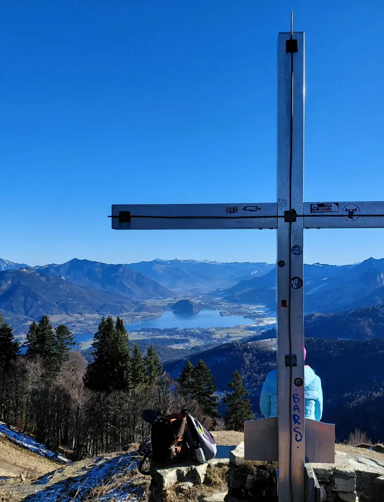Eine panoramische Aussicht von einem Berggipfel mit einer Person, die neben einem Kreuz sitzt. Berge, ein See und eine Stadt sind in der Ferne unter einem klaren blauen Himmel zu sehen.