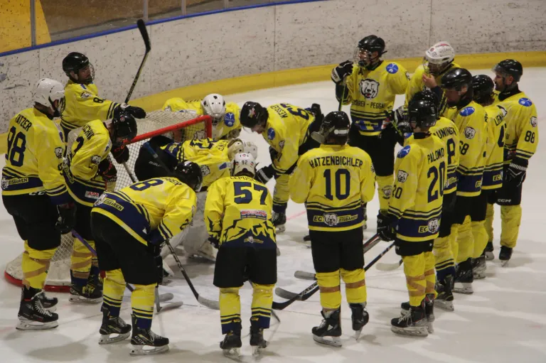 Eine Eishockeymannschaft in gelben Trikots drängt sich um ihr Tor. Die Spieler tragen Helme und Eislaufschuhe. Die Nummern auf ihren Trikots reichen von 8 bis 20. Ein Spieler beugt sich nach unten, während andere umherschauen.