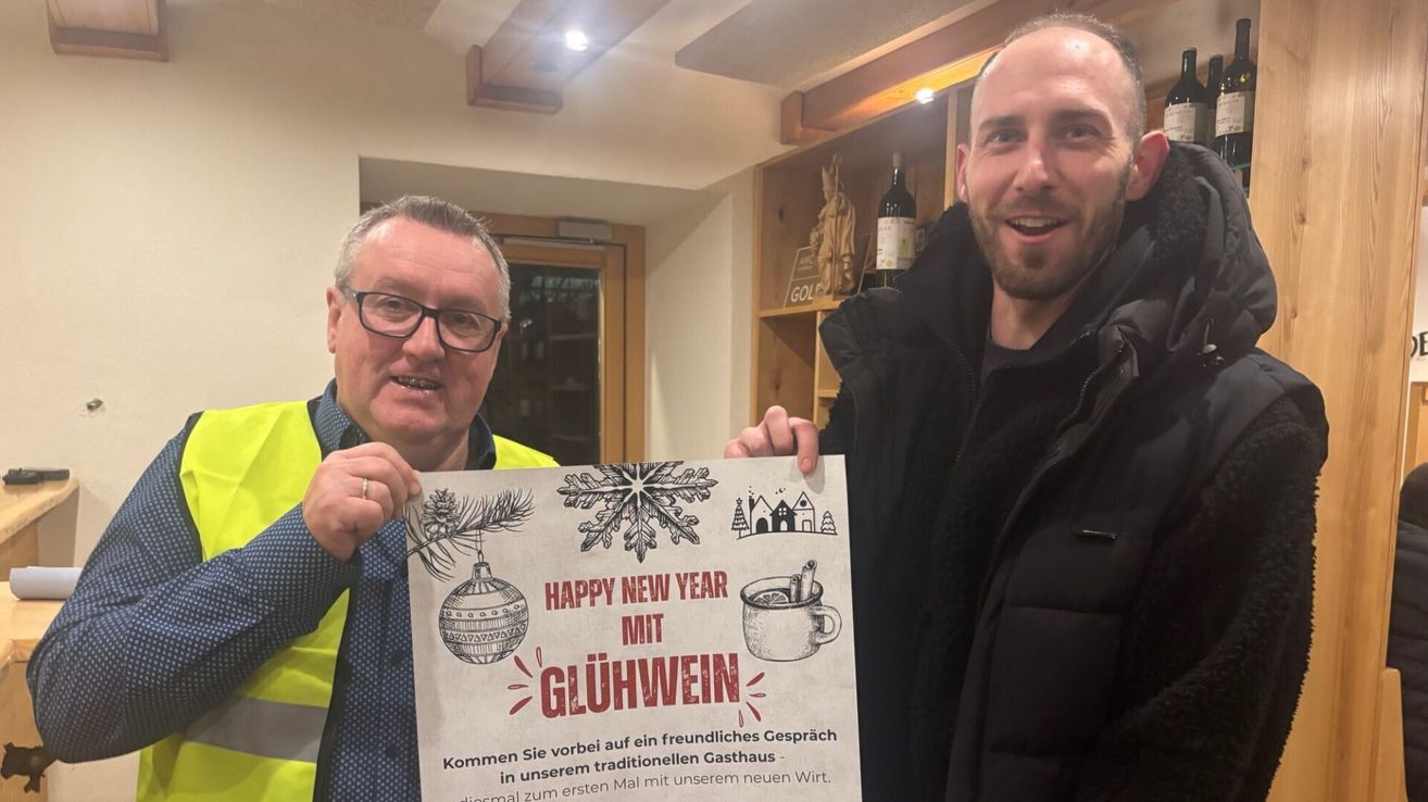 Zwei Männer halten ein Schild mit 'Frohes Neues Jahr mit Glühwein'. Sie lächeln und scheinen sich auf einer Party oder einem Treffen zu befinden.