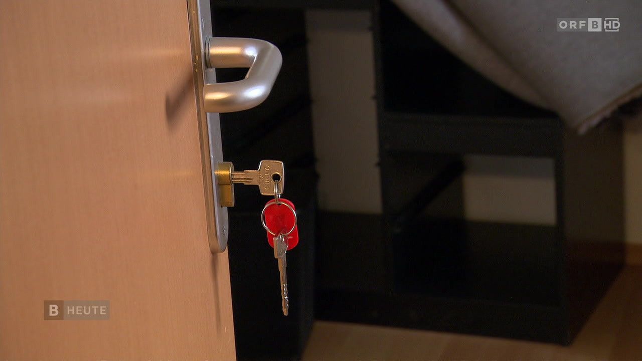 Ein Nahaufnahme einer Türgriff mit eingelegtem rotem Schlüsselbund. Der Schlüsselbund besteht aus zwei Schlüsseln und einem kleinen roten Gegenstand. Hinter der Tür befindet sich eine verschwommene Treppe.