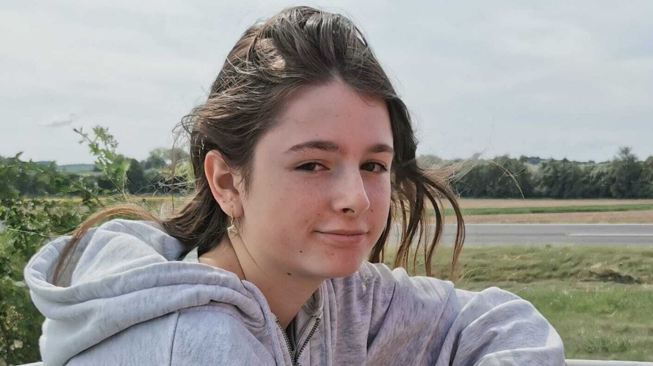 Eine Frau mit braunen Haaren in einer grauen Kapuzenjacke lächelt und steht auf einem Grasfeld mit Bäumen und einer Straße im Hintergrund.