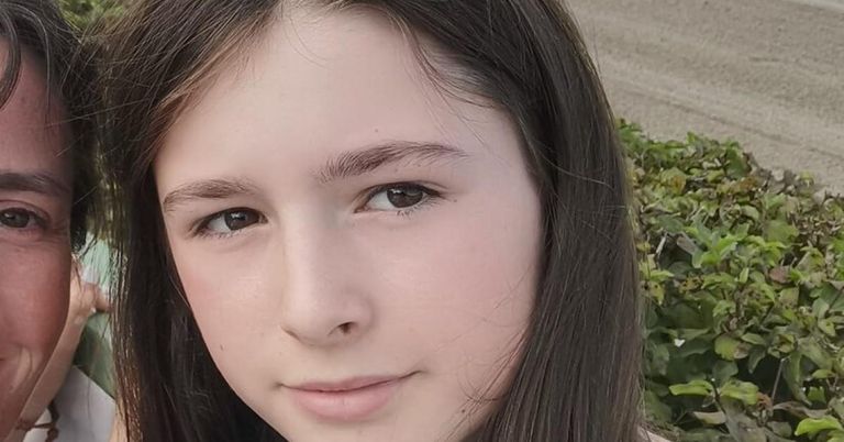 Ein junges Mädchen mit dunkelbraunen Haaren und Augen lächelt direkt in die Kamera. Ihr Gesicht ist leicht geneigt und ihre Lippen sind leicht geöffnet. Der Hintergrund ist verschwommen mit grünem Laub.